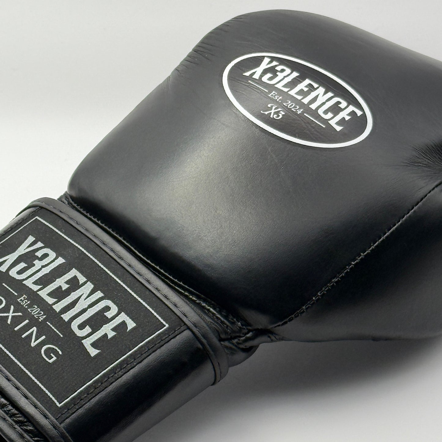 Close-up van zwarte X3LENCE bokshandschoen met wit logo en stevige klittenbandsluiting – echt leer, hoogwaardige afwerking, beschikbaar in 10 oz, 12 oz, 14 oz en 16 oz.