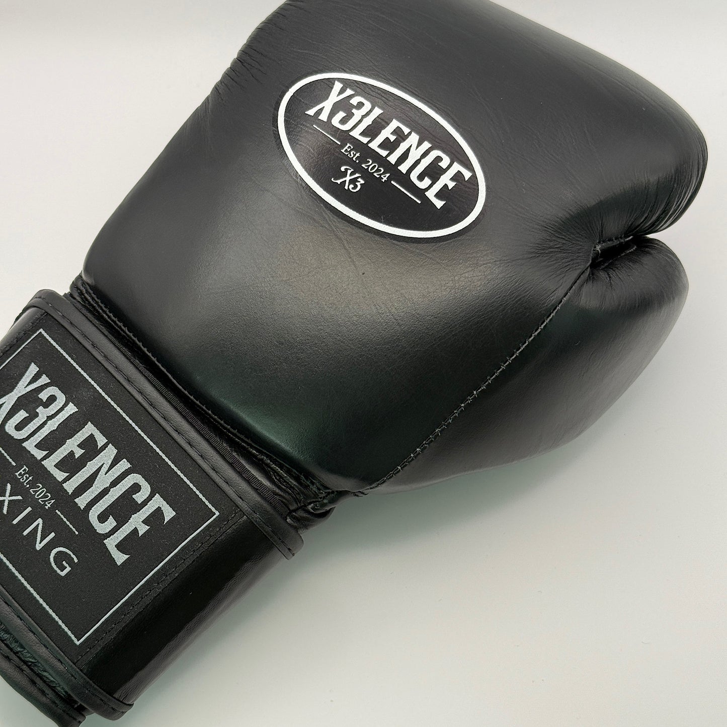 Zwarte X3LENCE bokshandschoen in close-up – minimalistisch en krachtig ontwerp met contrasterend wit logo, gemaakt van echt leer voor duurzaamheid en professionele uitstraling. Verkrijgbaar in 10 oz, 12 oz, 14 oz en 16 oz.